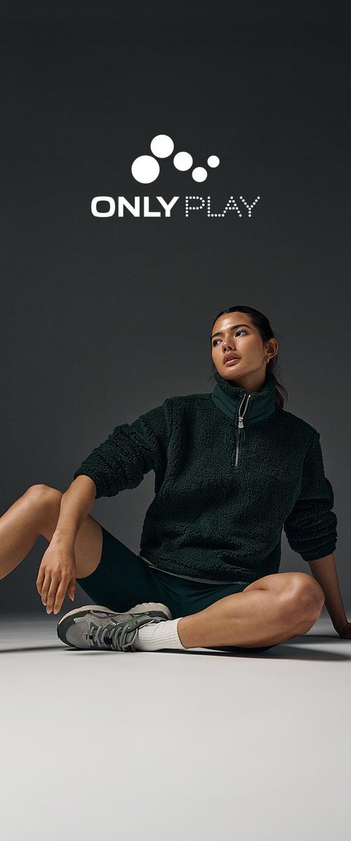 Femme en veste polaire vert foncé, short noir, chaussettes blanches et baskets grises assise par terre devant un fond sombre avec le logo "Only Play" au-dessus.