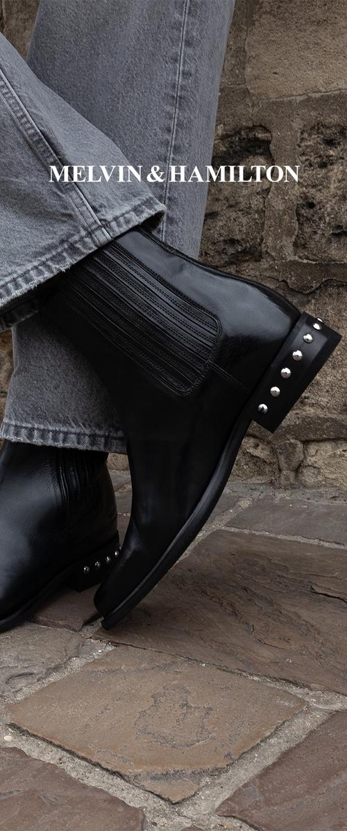 Bota corta de cuero negro con elástico acanalado en los costados, punta redonda y tachuelas plateadas decorativas en el talón. Usada con vaqueros grises.
