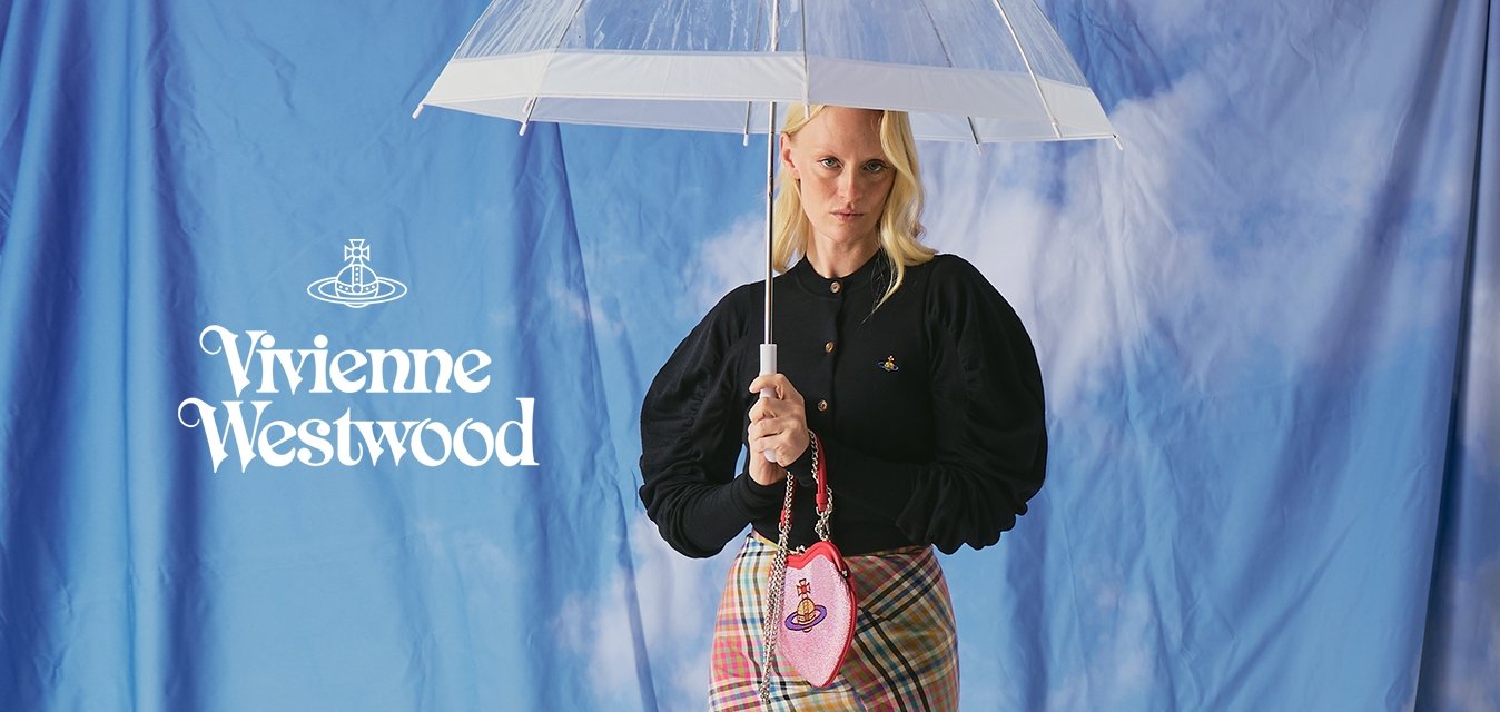 Blond žena v černém topu s nafouklými rukávy a kostkované sukni drží průhledný deštník a růžovou kabelku na pozadí modré oblohy s mraky.