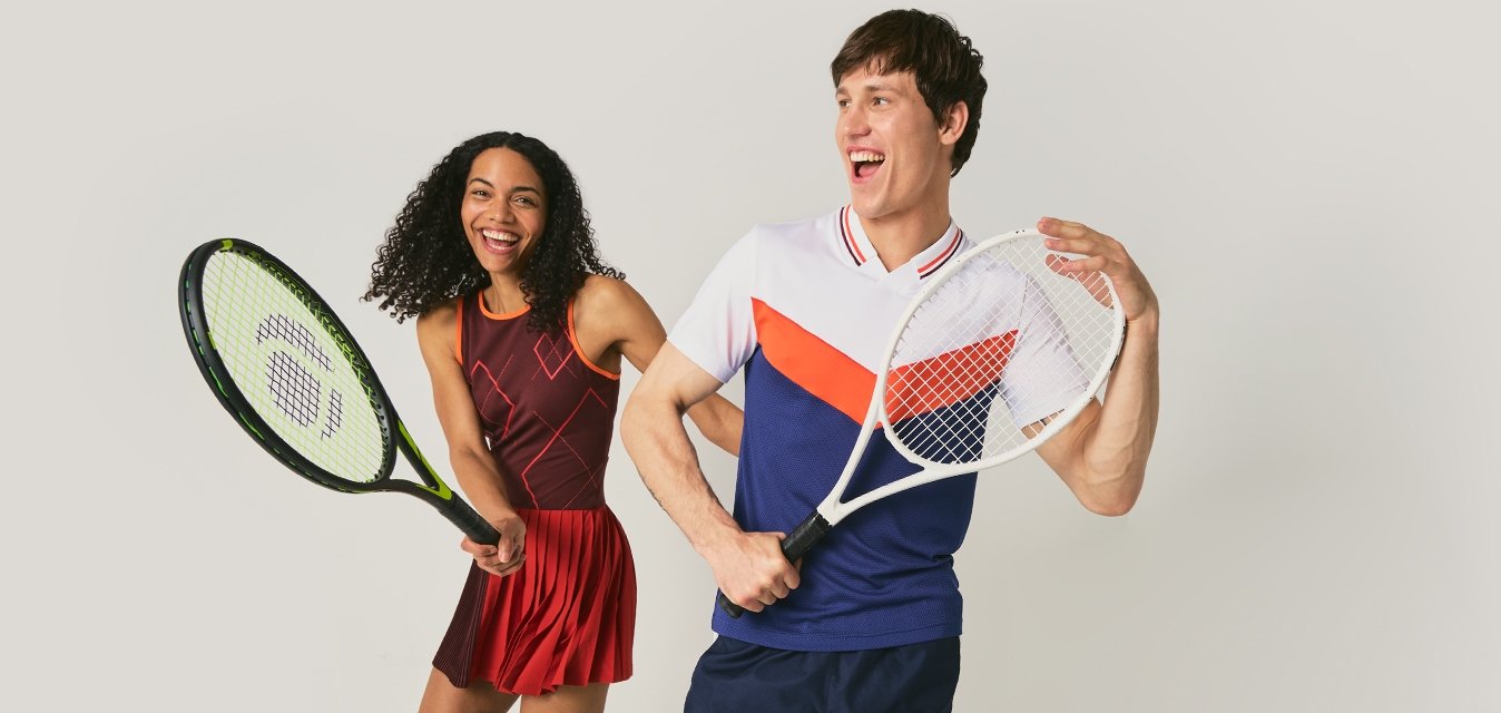 Usměvavý muž a žena v tenisovém oblečení pózují s raketami, stojí před jednoduchým světlým pozadím.