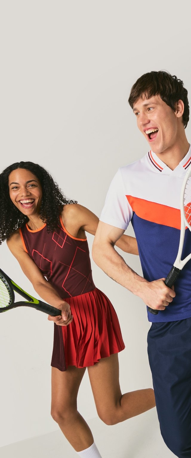 Tennisoutfit: vrouw in een nauwsluitende, kastanjebruine jurk met geplooide rok, man in een wit polo met blauwe en oranje accenten; beiden houden rackets vast.
