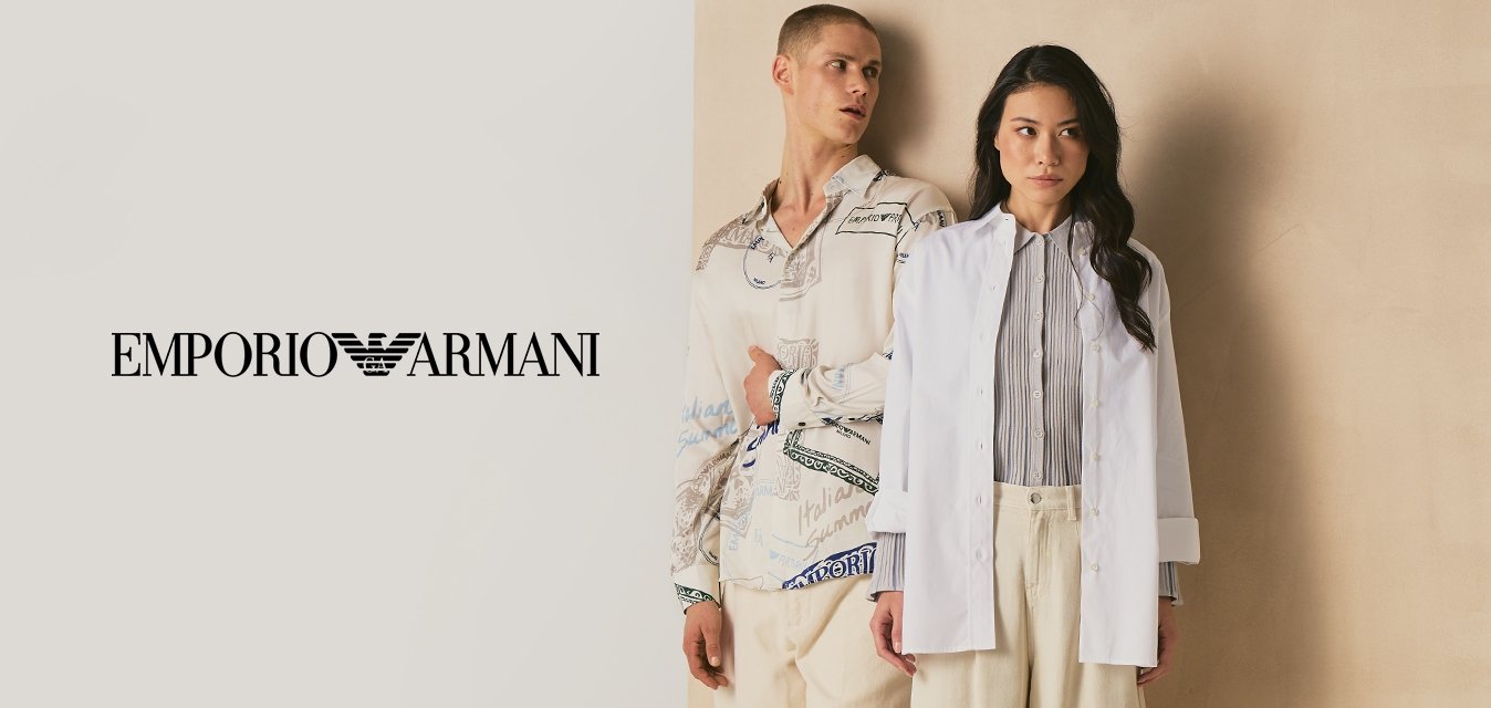 Dvě osoby stojí před béžovým panelem, jedna má na sobě vzorovanou košili a krémové kalhoty, druhá bílý blejzr a volné krémové kalhoty.