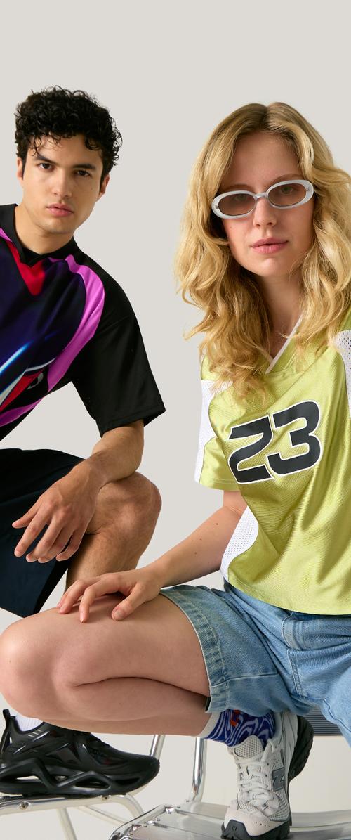Joven hombre y mujer agachados, vestidos con ropa deportiva informal, la mujer con gafas de sol y pantalones cortos vaqueros, el hombre con una camisa colorida y pantalones cortos negros.