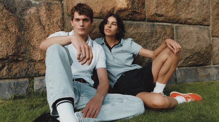 Zwei Personen sitzen auf dem Gras gegen eine Steinwand. Der Mann trägt ein weißes Polohemd und helle Jeans; die Frau trägt ein hellblaues Polohemd und schwarze Shorts.