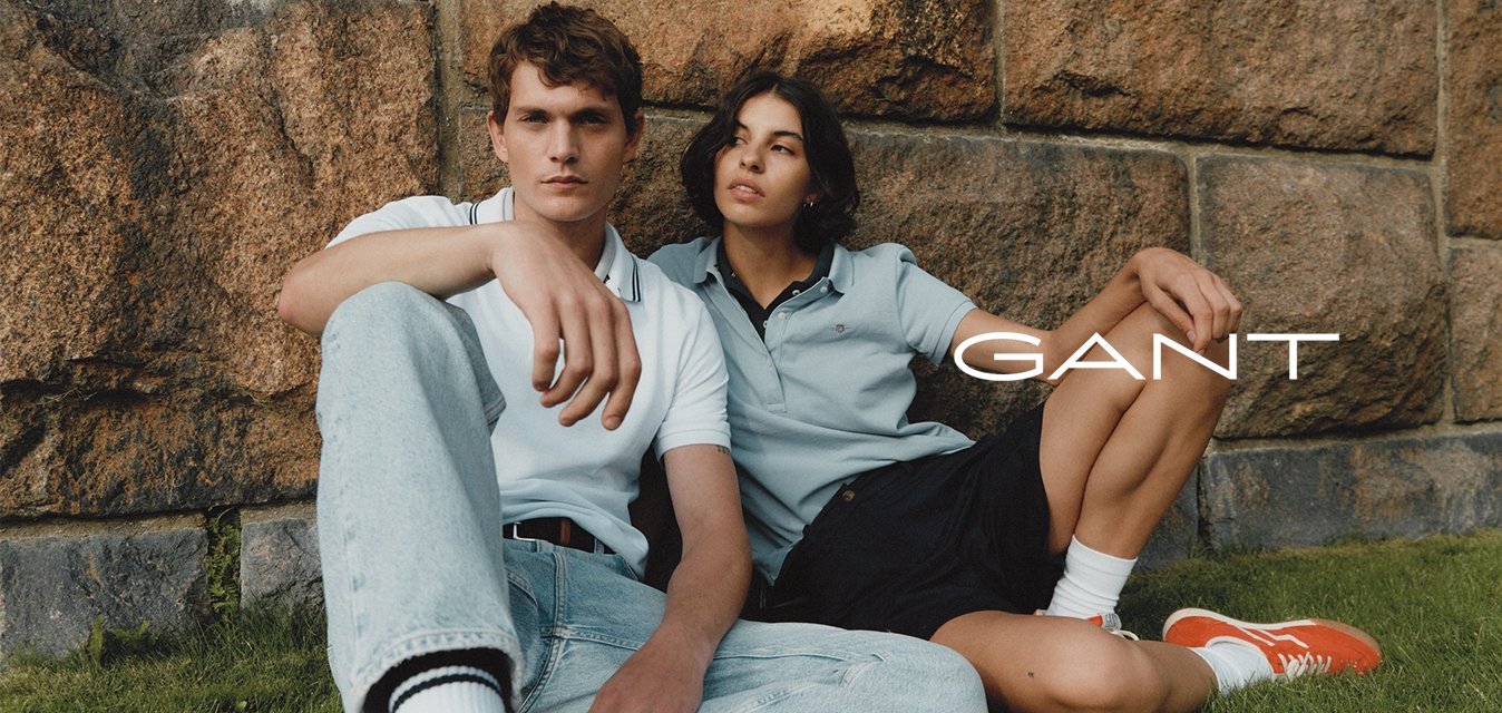 Dva jednotlivci sedí u kamenné zdi. Muž má na sobě bílé polo tričko a světlé modré džíny; žena má na sobě světlé modré polo tričko a černé šortky.