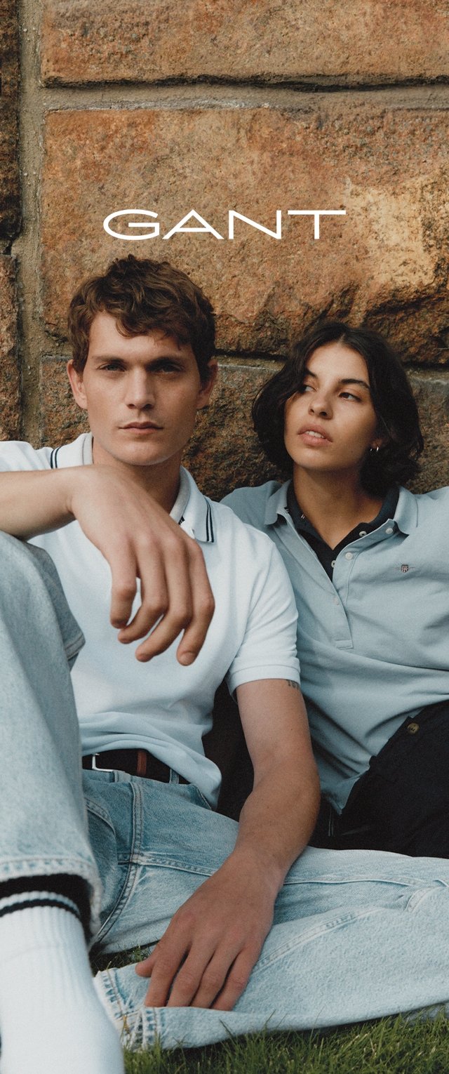 Två personer sitter på gräs mot en stenbakgrund. Mannen har på sig en vit polo med svarta accenter och ljusblå jeans. Kvinnan har på sig en ljusgrå polo.