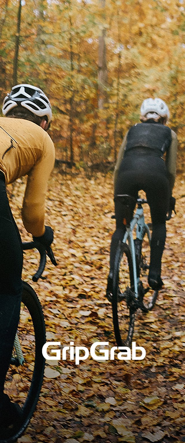 Deux cyclistes roulent sur un chemin recouvert de feuilles. L'un porte un maillot à manches longues brun, l'autre est en noir, tous deux sur des vélos de gravel.