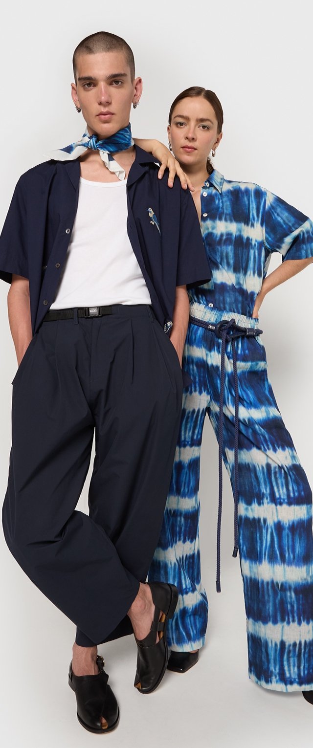 Deux modèles se tiennent côte à côte. À gauche, un homme porte une chemise courte à manches navy, un T-shirt blanc et un pantalon navy plissé. À droite, une femme porte une combinaison tie-dye bleue et blanche avec une taille nouée et des manches courtes. Tous deux portent des chaussures noires.