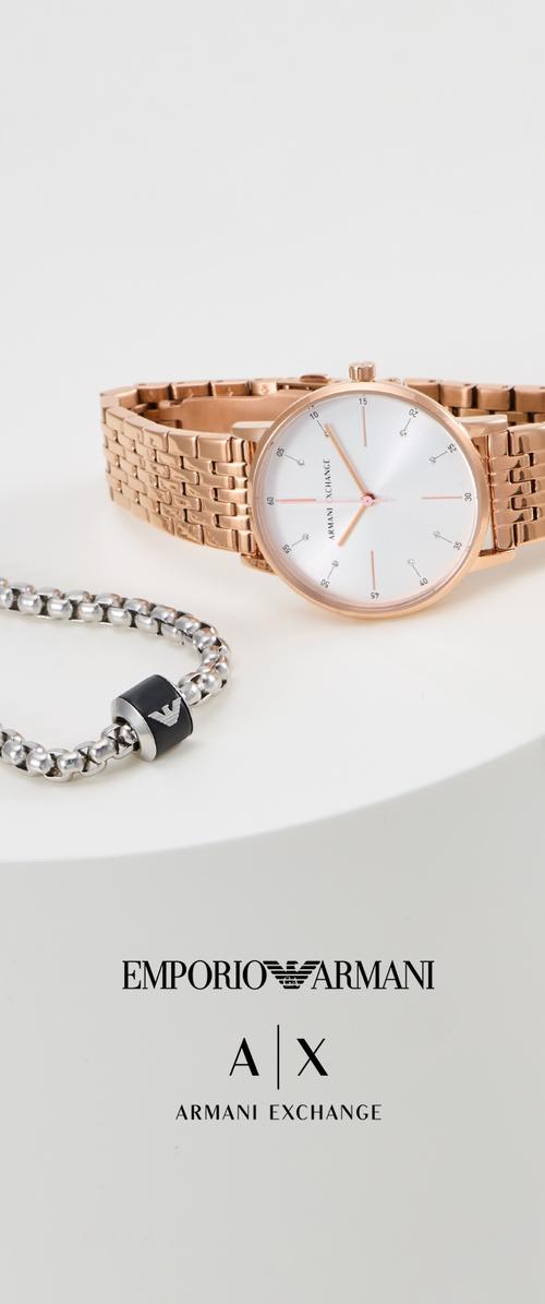Reloj de oro rosa con una correa de metal texturizado, esfera blanca y acentos plateados, acompañado de una pulsera de cadena plateada con un emblema negro.