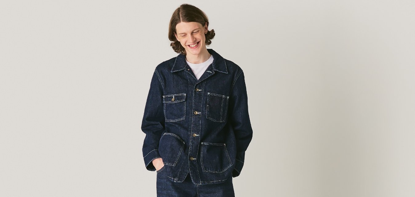 Osoba v tmavé džínové bundě a kalhotách stojící s rukama v kapsách na světle šedém minimalistickém pozadí.