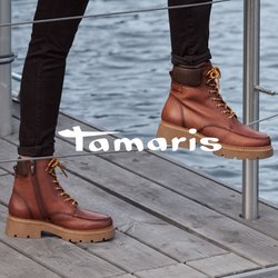 Hnědý nepromokavý kabát s límcem, krémový svetr, černé kalhoty a hnědé kotníkové boty s tkaničkami a silnou podrážkou. Dřevěné a kovové prostředí.