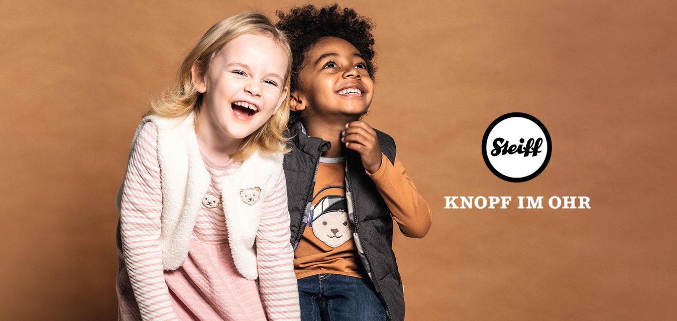 Dvě děti sedí na krabici. Dívka má na sobě růžové pruhované šaty a krémovou vestu; chlapec má na sobě šedou vestu přes tričko s grafickým potiskem hnědého medvěda a džíny.