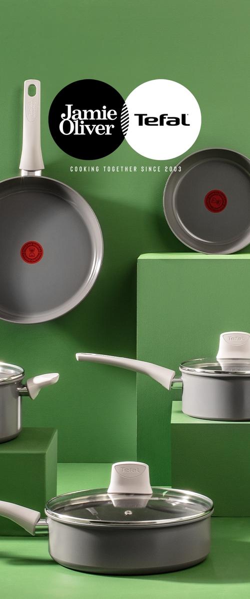 Graue Antihaft-Pfannen und -Töpfe mit weißen Griffen, präsentiert auf grünen Blöcken, mit Jamie Oliver und Tefal Logos und dem Slogan über das Kochen seit 2003.