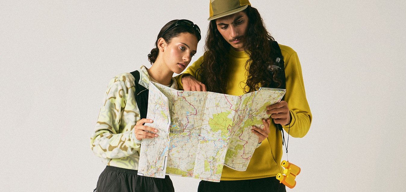 Dva turisté v kraťasech a teniskách, jeden v žluté čepici, oba studují složenou mapu na jednobarevném pozadí.