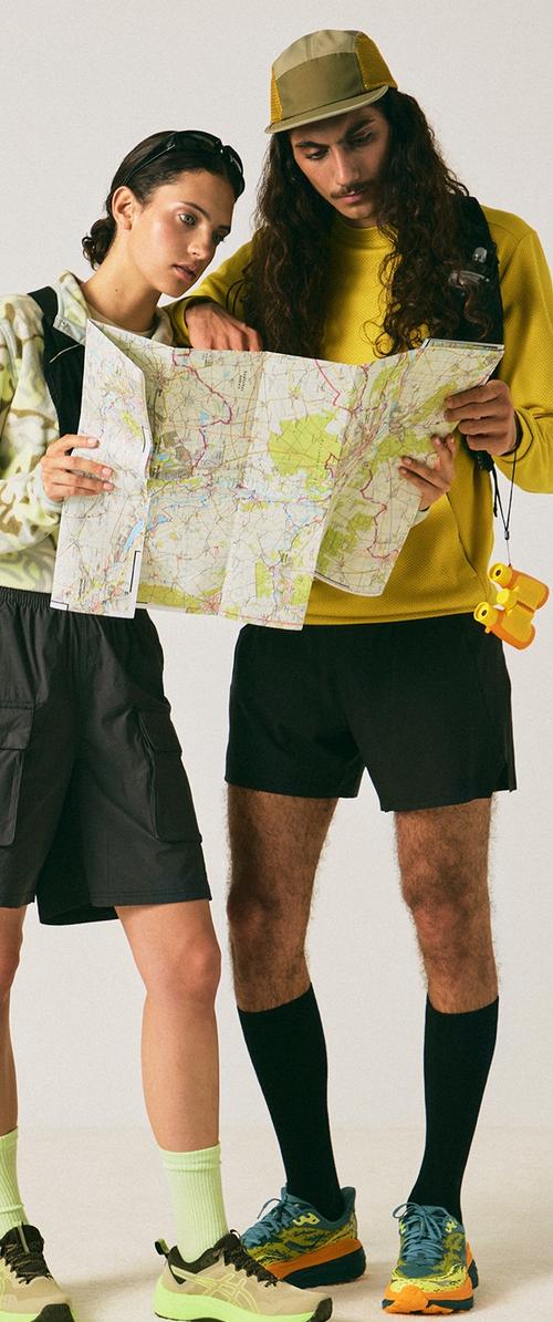 Dvaja turisti s batohmi študujú veľkú zloženú mapu; jeden drží žlté ďalekohľady, obaja sú oblečení v neformálnom outdoor oblečení a športovej obuvi.