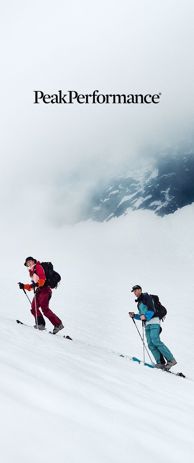 Zwei Skifahrer in bunter Ausrüstung steigen mit Rucksäcken und Skistöcken einen verschneiten Berghang unter einem nebligen Himmel hinauf.