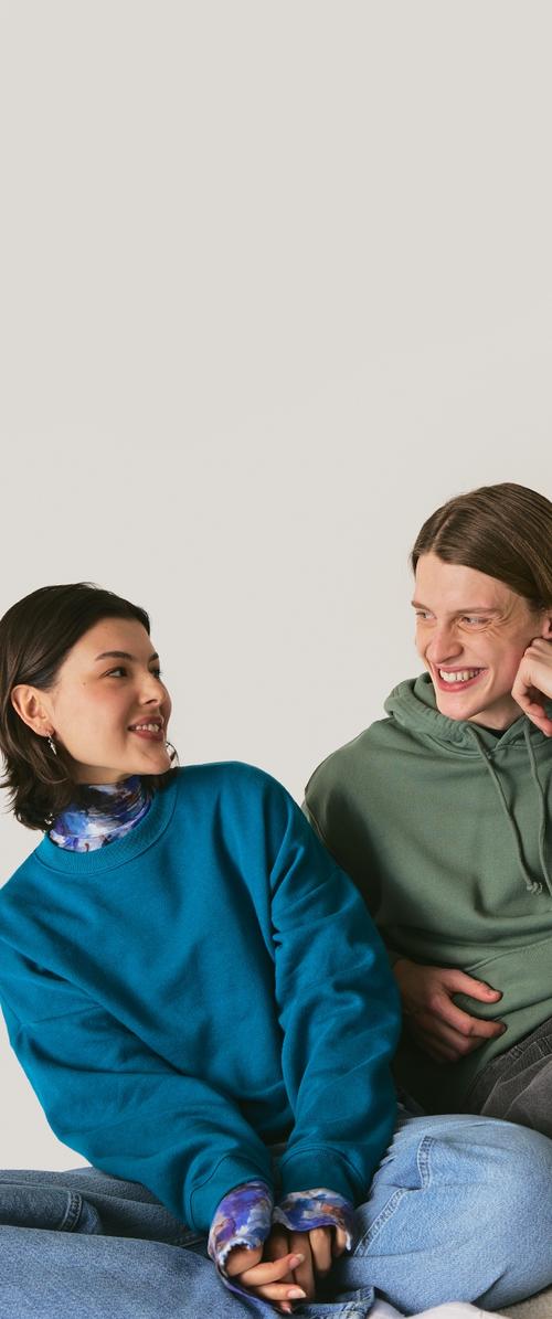 Två personer sitter nära varandra, klädda i en teal-färgad sweatshirt och en grön hoodie, tillsammans med blå jeans. Teal-färgen har en texturerad yta.