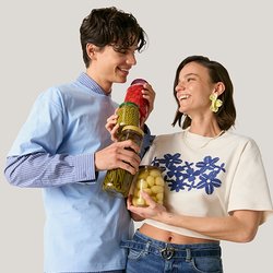 Mladý muž a žena se usmívají, drží sklenice s nakládanou zeleninou a stojí před jednobarevným světlým pozadím.