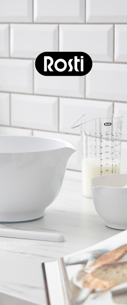 Ciotole bianche per mescolare e brocca dosatrice con latte sul piano cucina contro parete piastrellata bianca, con logo Rosti sopra.