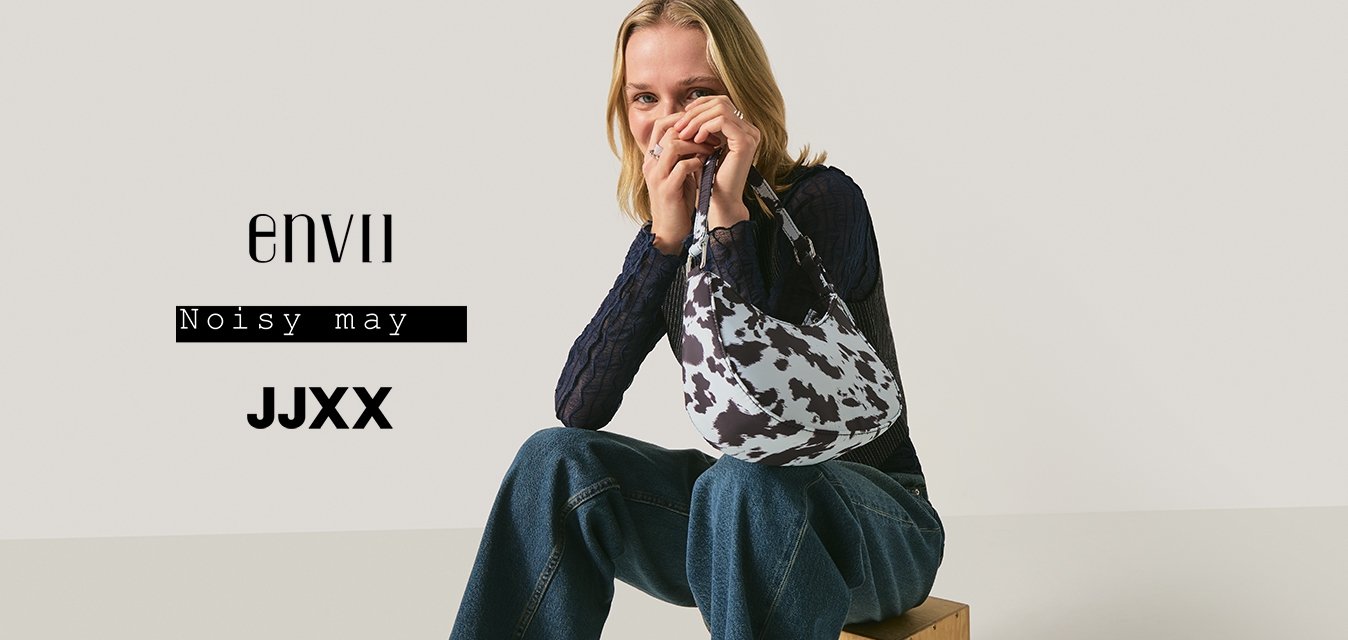 Mladá žena sedící na dřevěné bedně, držící kabelku s černobílým vzorem kraví kůže, oblečená v námořnicky modrém krajkovém topu a širokých džínách.