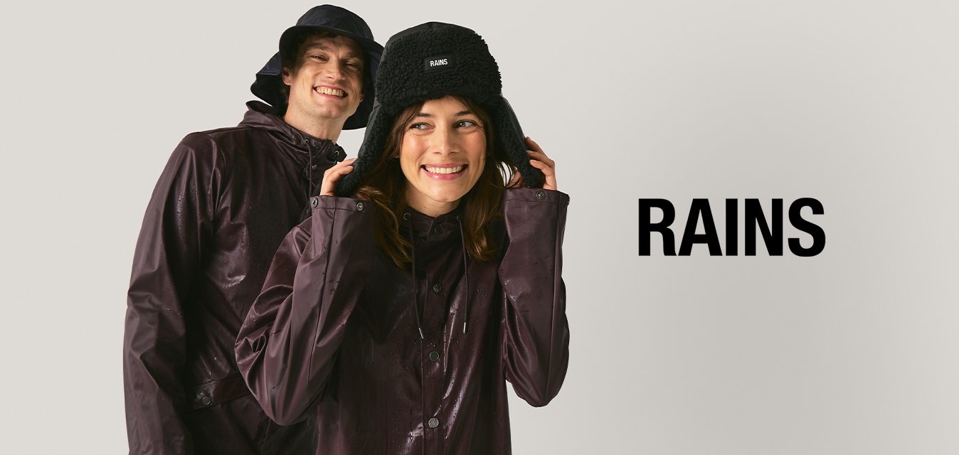 Dva lidé v tmavě fialových nepromokavých bundách a černých kloboucích se usmívají na jednobarevném světlém pozadí.