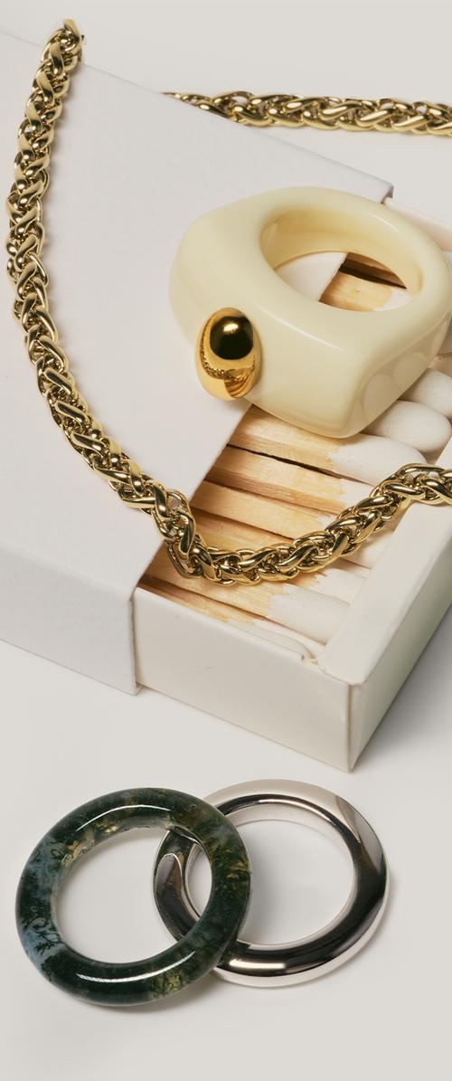 Guld kæde ligger på en åben hvid tændstiksæske med en cremefarvet ring, der har en guld accent; mørkegrønne og sølvfarvede cirkulære ringe ligger i nærheden på en hvid overflade.