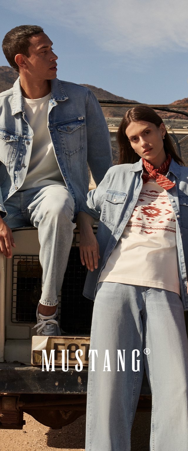 Hombre y mujer con ropa vaquera sentados y apoyados en un vehículo al aire libre, con colinas desérticas al fondo.