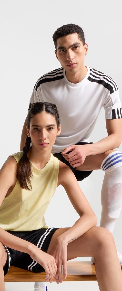 Deux modèles posent en vêtements de sport : un débardeur jaune et un short noir avec des rayures blanches ; une chemise de sport blanche et un short noir avec des accents bleus.