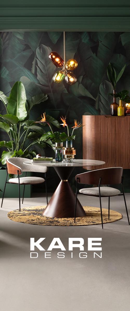 Mesa redonda de mármol blanco con base de madera cónica, dos sillas tapizadas, plantas tropicales, armario de madera y lámpara colgante.