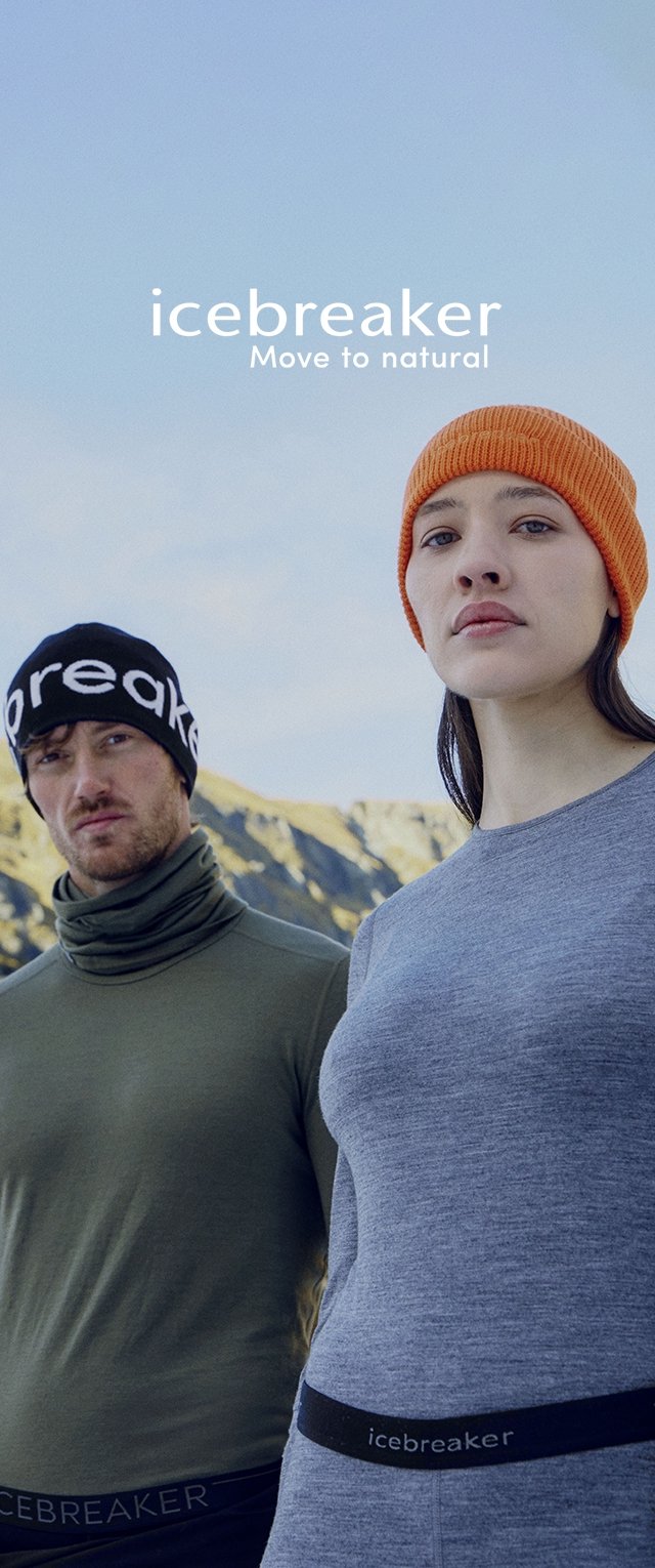 Dos personas llevan ropa para exteriores de Icebreaker. Una lleva una camiseta gris con una pretina negra con la marca, y la otra lleva una camiseta verde con un pasamontañas negro, ambas bajo un cielo azul.