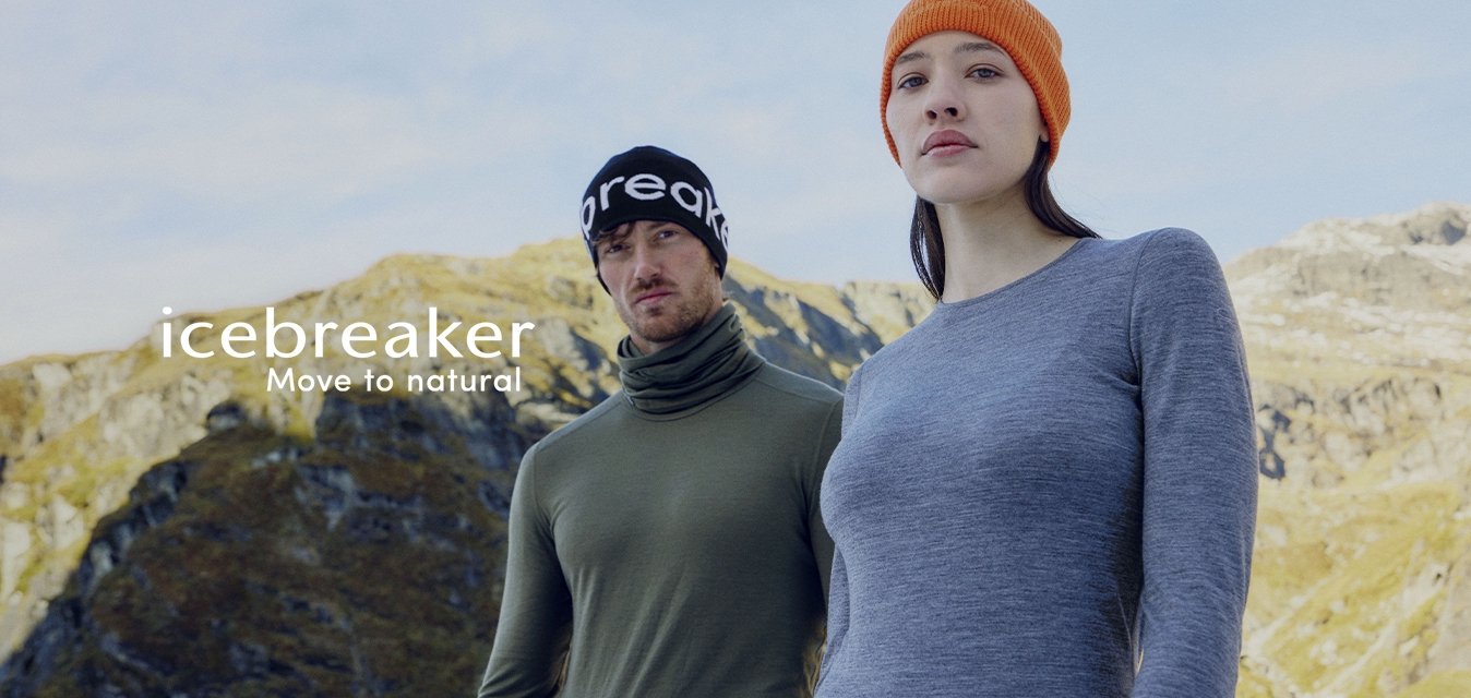 Dva dospělí v outdoorovém oblečení s potiskem značky na spodním prádle a čepicích stojící před horským terénem pod jasnou oblohou.