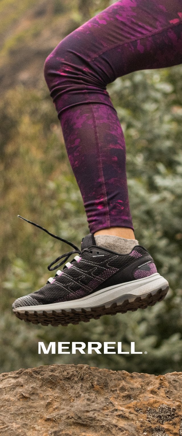 Zapatilla deportiva Merrell en negro y morado con una suela texturizada, suspendida en el aire contra un fondo natural, acompañada de leggins morados con patrón.