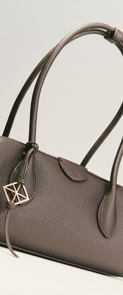 Sac à main en cuir brun foncé avec deux anses et un pendentif métallique géométrique sur une surface texturée sur fond uni.