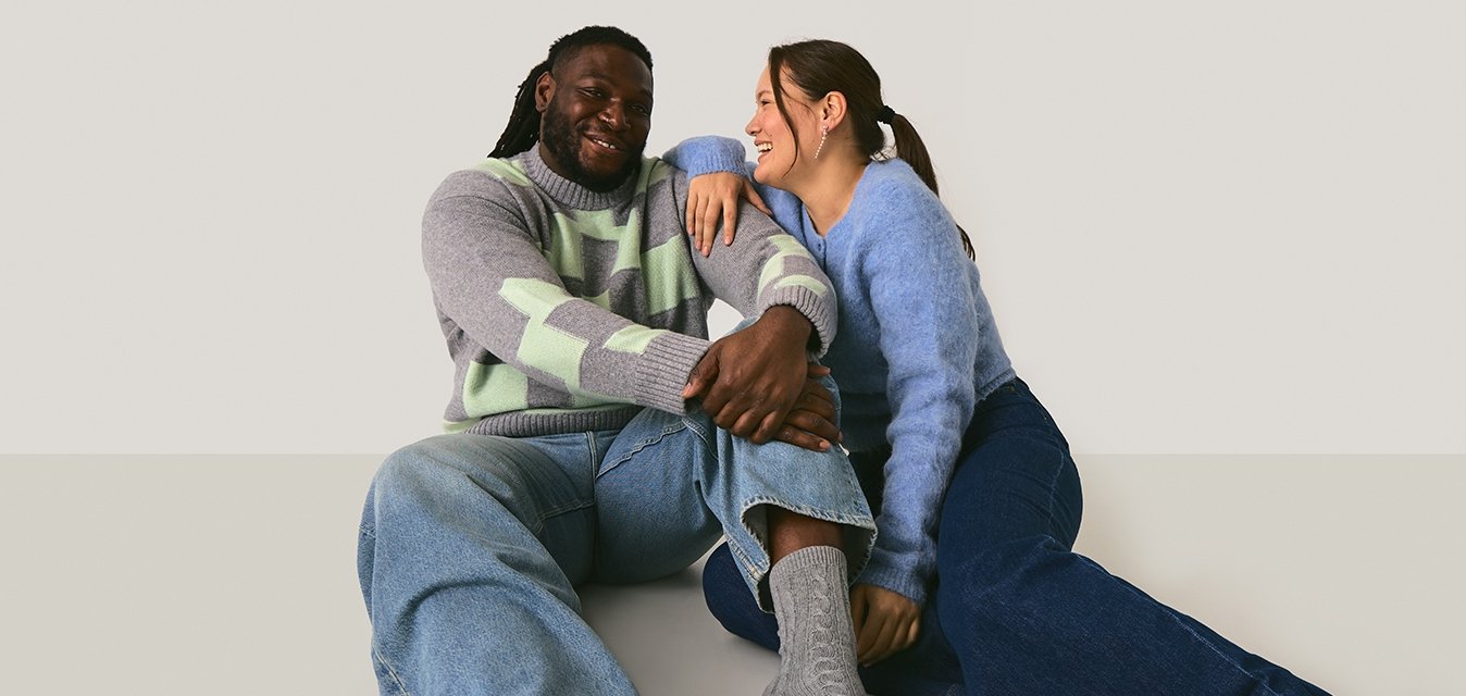 Pletené svetry v světle šedé a modré barvě s zelenými akcenty, volné džínsy a vzorované šedé ponožky, sedící na světlém pozadí.