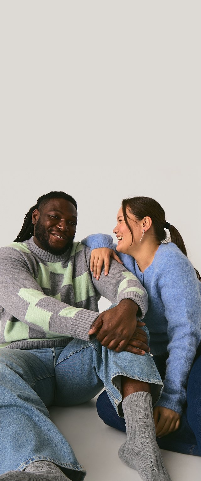 Zwei Personen sitzen zusammen. Eine trägt einen grauen Pullover mit grünen geometrischen Mustern, die andere einen blauen, flauschigen Pullover. Sie tragen Jeans und gemusterte Socken.