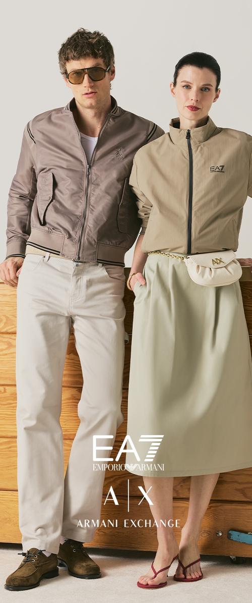 Hombre con chaqueta bomber marrón claro y pantalones blancos con gafas de sol, mujer con chaqueta beige con cremallera, falda midi y riñonera blanca, de pie uno junto al otro.