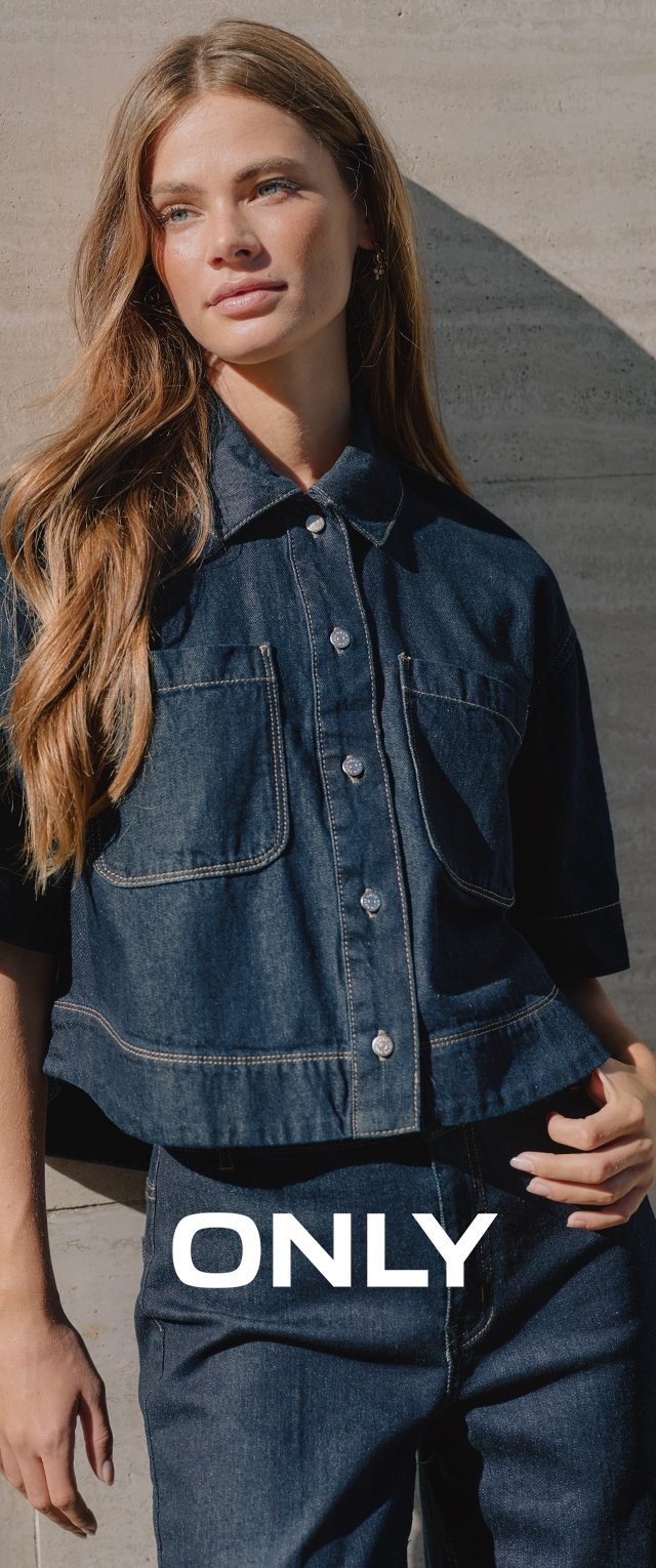 Junge Frau mit langen Haaren, trägt eine dunkle Denimjacke und Jeans, steht vor einer sonnenbeschienenen Betonwand mit dem Schriftzug "ONLY" auf Taillenhöhe.