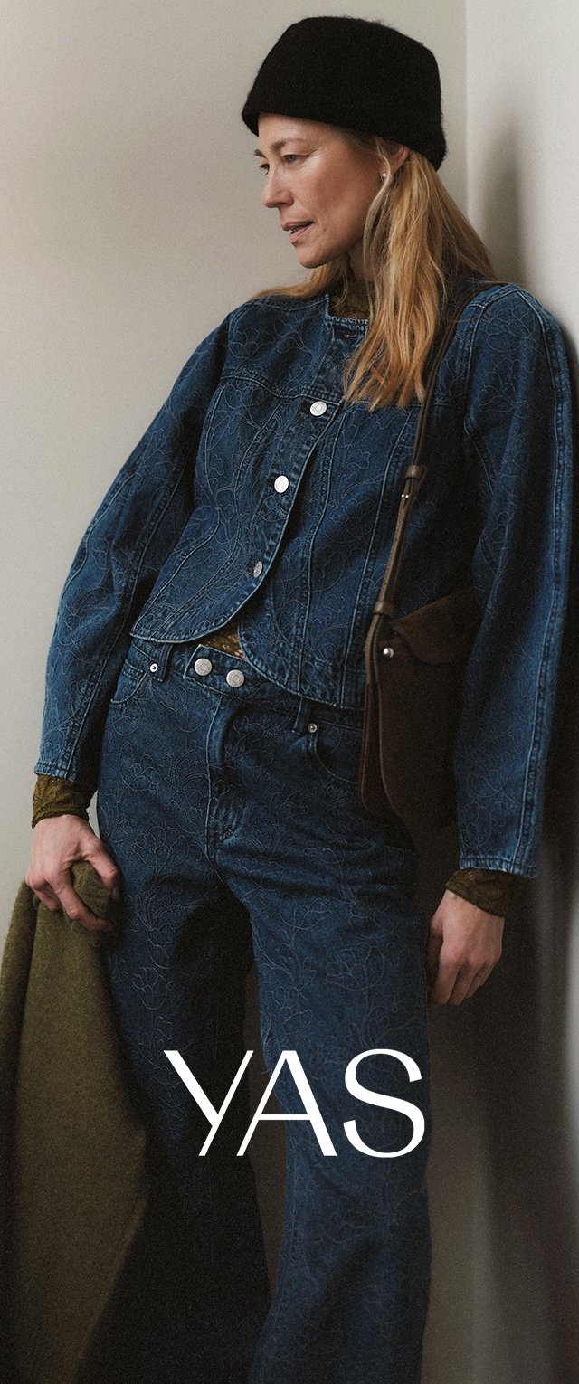 Femme portant une veste et un pantalon en denim à motifs, bonnet noir, sac à bandoulière marron, tenant un manteau vert, debout devant un fond uni.
