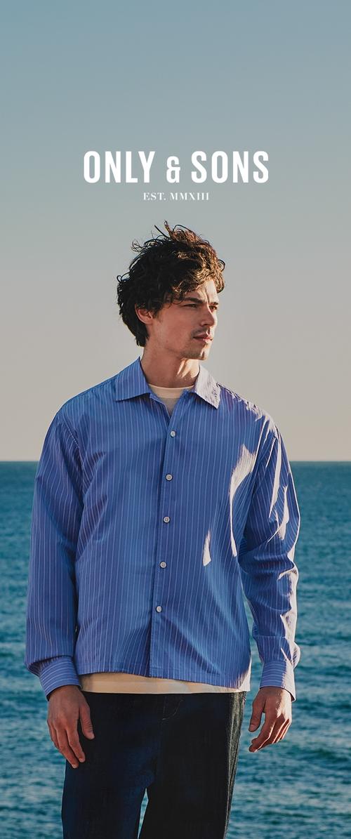 Hombre con cabello rizado vestido con una camisa de rayas azules y abotonada, de pie junto al océano bajo un cielo despejado, con el texto "ONLY & SONS EST. MMXIII" arriba.