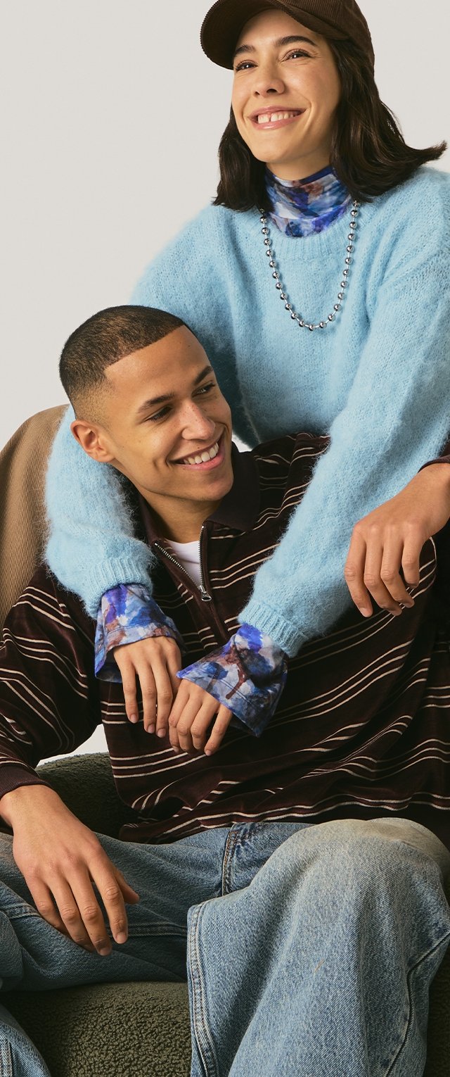 Dos personas sentadas juntas: una con un suéter peludo azul claro y la otra con una camiseta de rayas oscura; ambas llevan vaqueros cómodos.