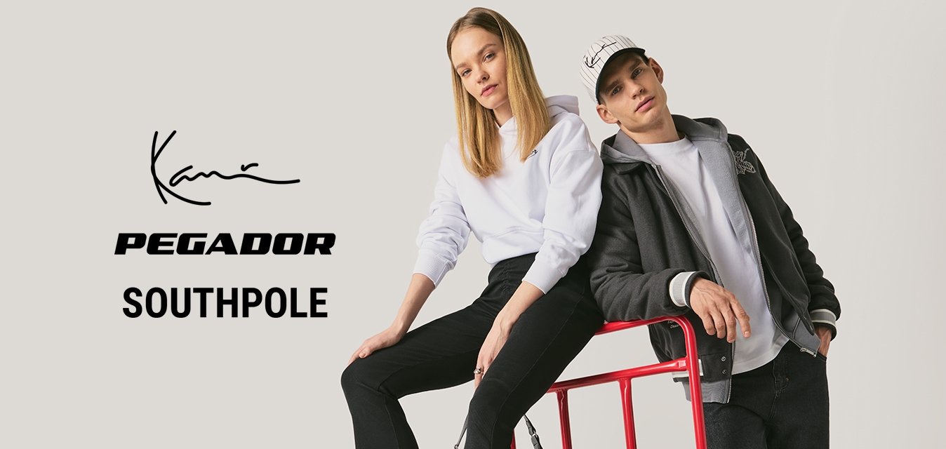 Žena v bílé mikině sedí na červeném plošinovém vozíku, zatímco muž v černé bundě a kšiltovce se opírá o jeho madlo v ateliérovém prostředí.