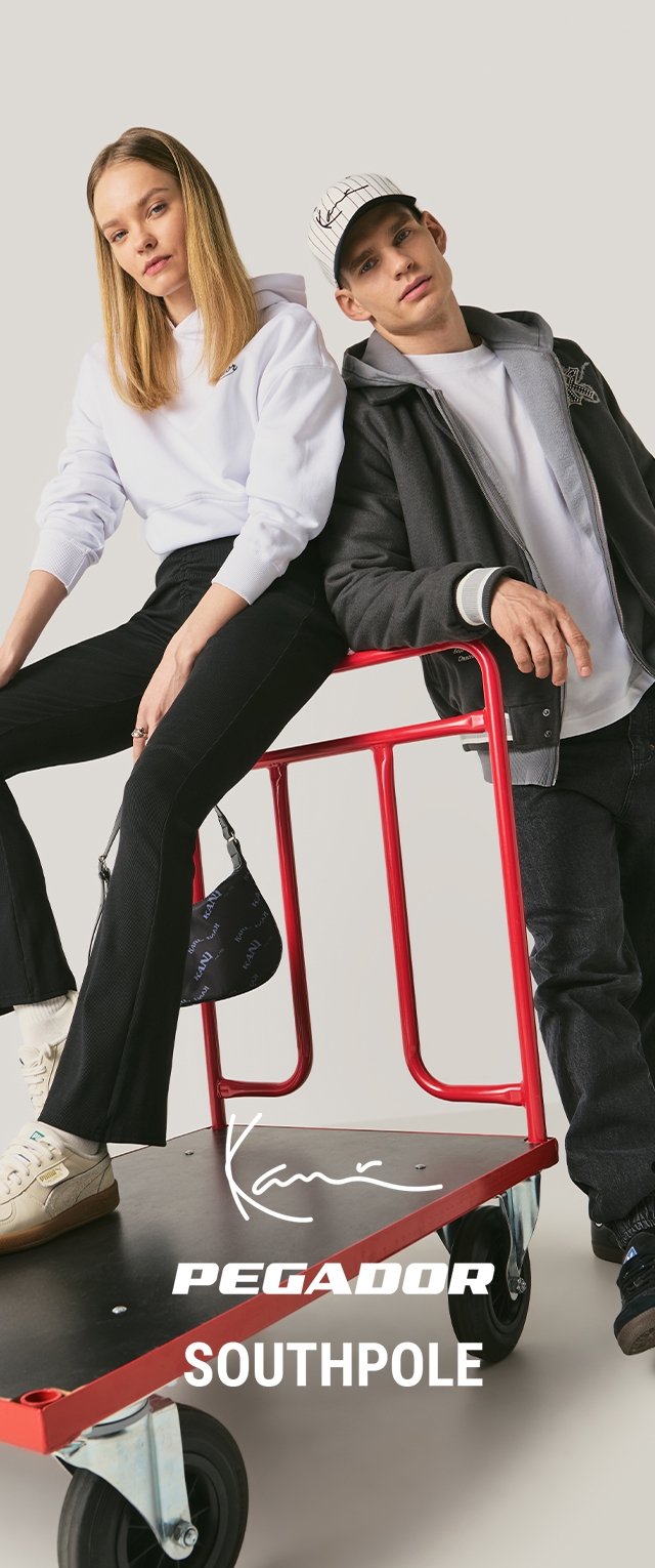 Zwei junge Erwachsene, eine Frau in weißem Kapuzenpullover und schwarzer Hose sitzt auf einem roten Plattformwagen, ein Mann in grauer Jacke und Kappe lehnt daran.