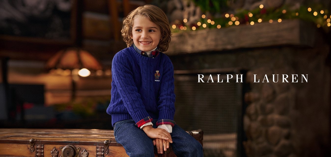 Usmívající se dítě s kudrnatými vlasy, oblečené v modrém svetr s pleteným vzorem a džínách, sedící na starožitné dřevěné truhle v útulné místnosti s kamenným krbem.