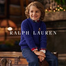 Usmívající se dítě s kudrnatými vlasy, oblečené v modrém svetr s pleteným vzorem a džínách, sedící na starožitné dřevěné truhle v útulné místnosti s kamenným krbem.