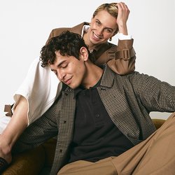 Dva modely sedí v hnědé židli. Jeden má na sobě kostkovanou košili, černý polo a béžové kalhoty; druhý je oblečen v hnědém kabátu přes bílé šaty.
