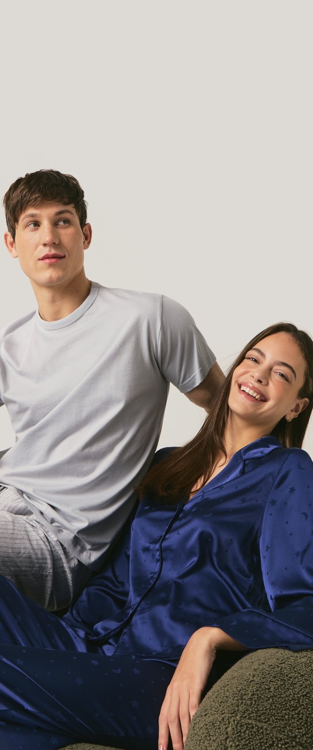 Joven con camiseta gris claro y pantalones a rayas junto a una mujer sonriente con pijama de seda azul oscuro sentada en una silla gris texturizada.