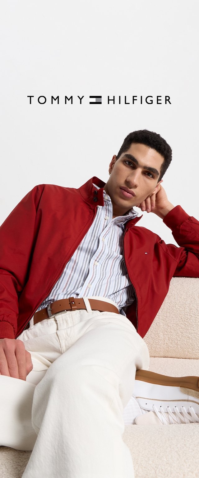 Veste zippée rouge sur une chemise rayée bleu et blanc, associée à un pantalon crème et des baskets blanches, assis sur une surface beige.
