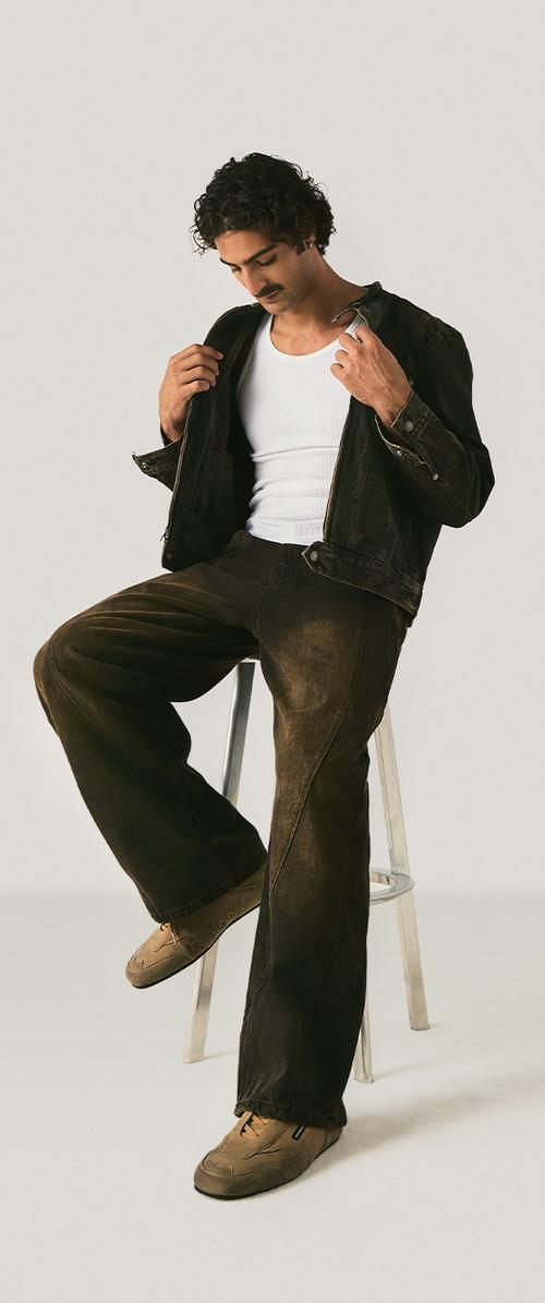 Mann mit dunklem lockigem Haar und Schnurrbart sitzt auf einem Hocker und richtet eine schwarze Jacke über einem weißen Tanktop, trägt braune Hose und beige Schuhe.