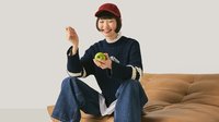 Junge Person mit roter Kappe und marineblauem Pullover, die auf einem beigefarbenen Ledersofa sitzt und lächelt, während sie einen grünen Apfel hält.