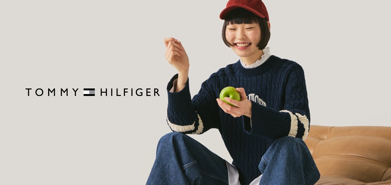 Mladá osoba v červené čepici, tmavě modrém svetru a džínách sedí na krémově hnědé kožené pohovce, drží zelené jablko a usmívá se na jednobarevném pozadí.